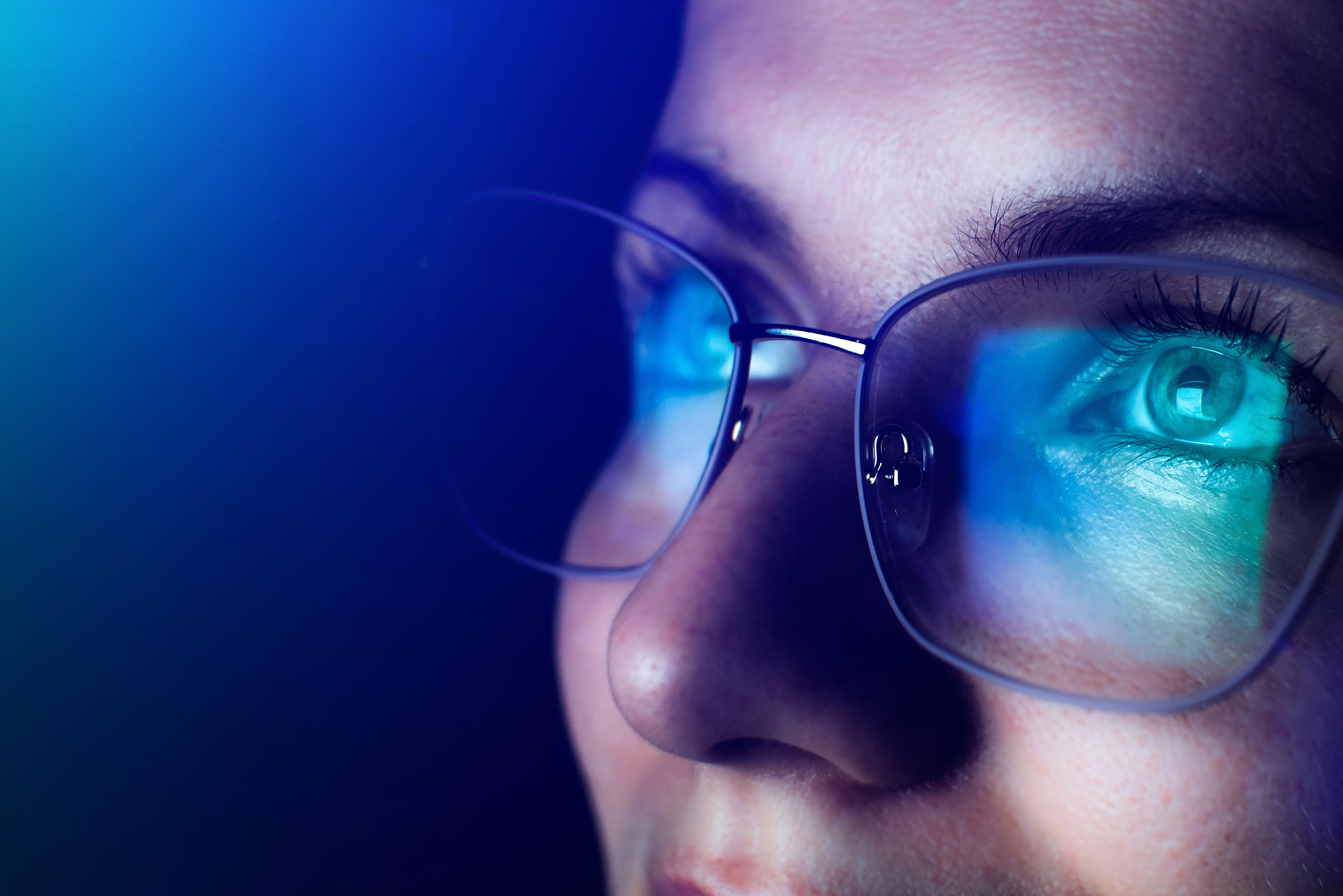 Close-up of a woman's face wearing glasses that reflect a bright blue screen, highlighting her focused eyes as she looks intently at content on a digital display.