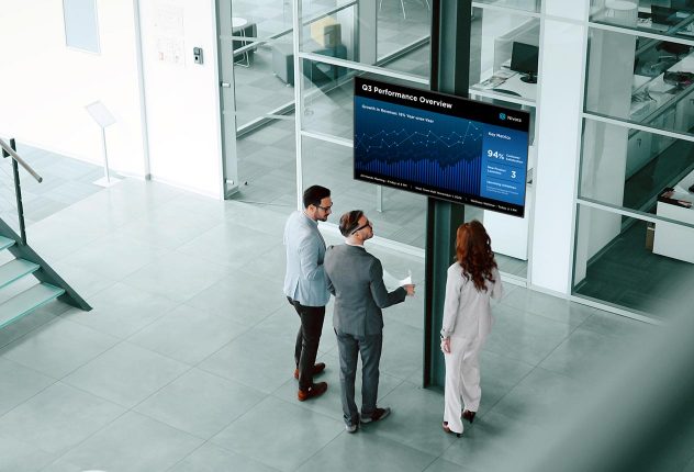 Three business professionals standing together in an open office space, analyzing data on a large digital screen labeled 'Q3 Performance Overview' with charts and metrics.