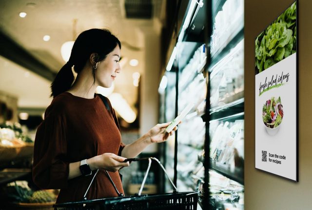 Woman in a grocery store checking a product in the refrigerated section, with a digital display showing a promotion for fresh salads and a QR code for recipes.