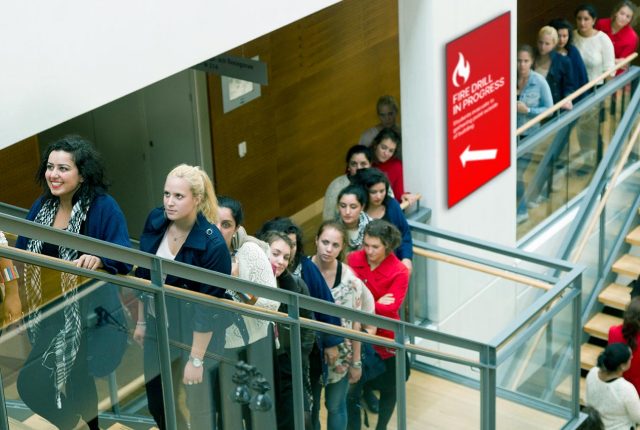 Large group of people moving in an orderly fashion up a staircase during a fire drill, with a digital display indicating 'Fire Drill in Progress' and an arrow pointing the way.