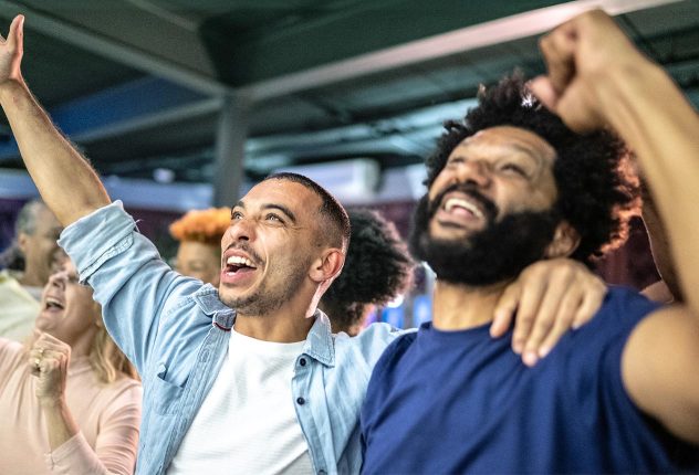 Two enthusiastic fans at a sports event cheering with raised arms, surrounded by other spectators showing excitement and joy.