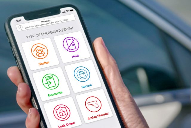 Hand holding a smartphone showing the Protect Alert application with icons for different types of events such as 'Shelter,' 'Evacuate,' 'Lock Down,' and 'Active Shooter.