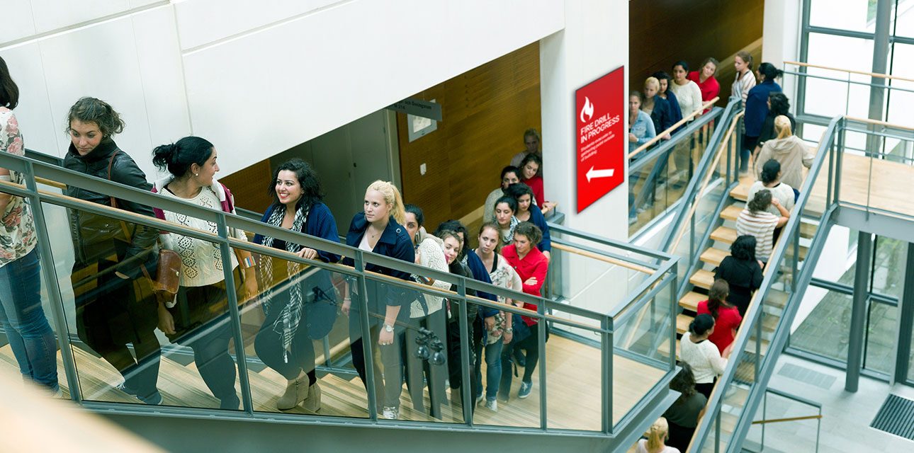 Large group of people moving in an orderly fashion up a staircase during a fire drill, with a digital display indicating 'Fire Drill in Progress' and an arrow pointing the way.